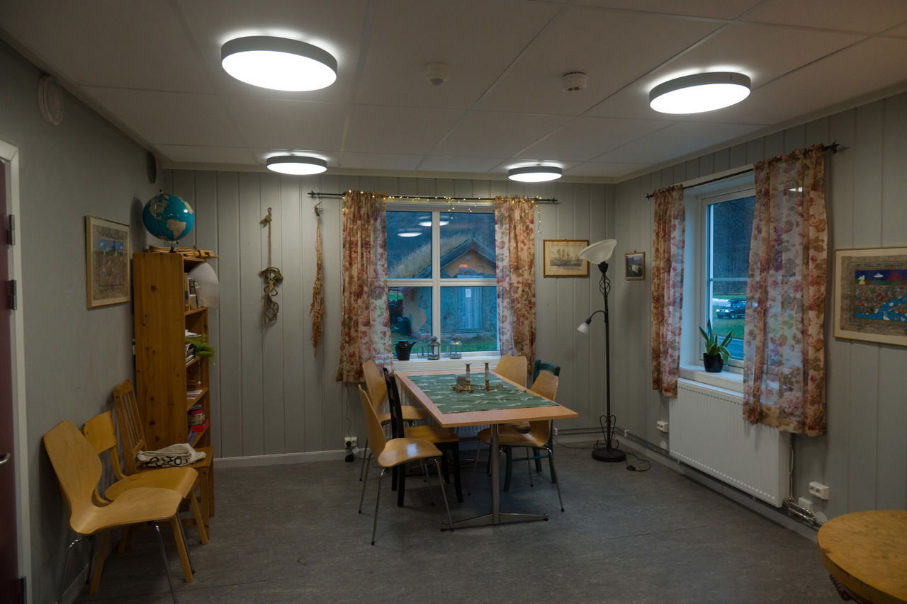 Fellesstove på internatet ved Sogn Jord- og Hagebruksskule, med spisebord, stolar, bokhylle og store vindauge som slepper inn naturlys.